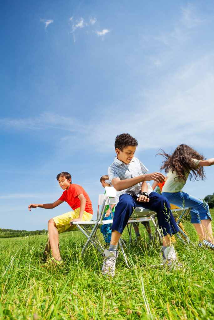 Kids playing game musical chairs and sitting fast on chairs in circle outside in summer