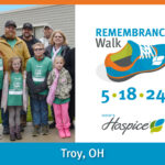 Remembrance Walk | 5/18/24 | Troy, OH Description: A group of smiling middle-aged people and children wearing fall coats stand grouped together for a posed photograph in front of a building. Most are wearing the 2023 Walk T-shirts.
